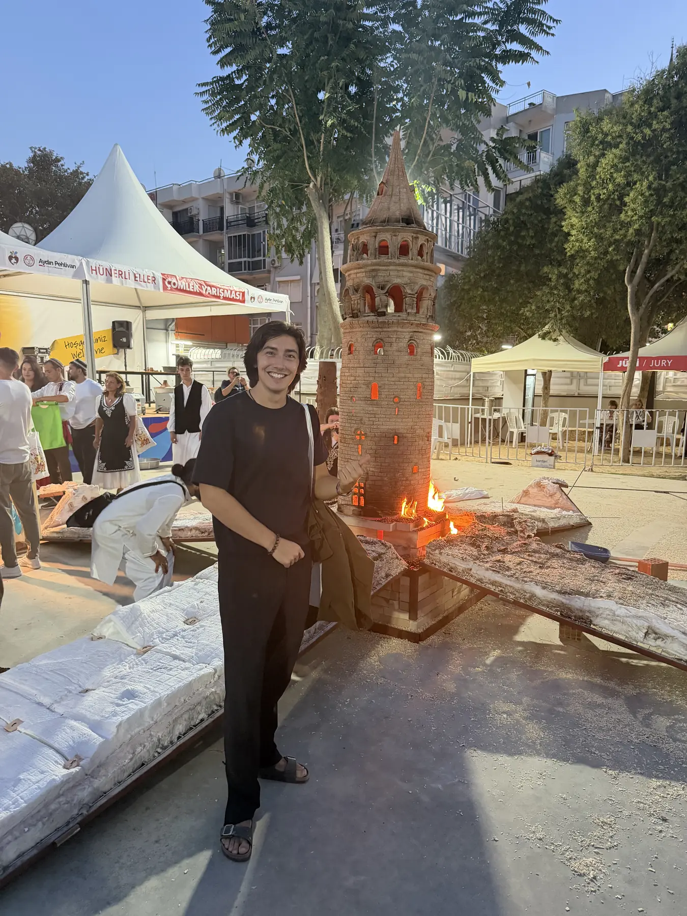 Daniel Pontoriero beside a terracotta tower with a fire at its base on the Menemen festival grounds, 2026
